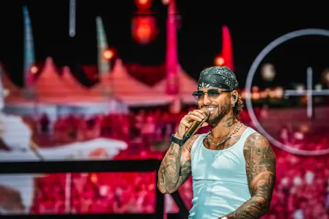 Maluma sonríe en camiseta de tirantes con el festival al fondo. Cook Music Fest, Tenerife, julio 2024.