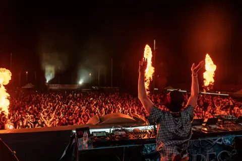 Fisher from behind, arms raised, before the crowd and the flames. Maudes Festival, Gran Canaria, July 2026.