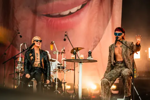 Ca7riel and Paco Amoroso seated on stage before a giant face. Granca Live Fest, Gran Canaria, July 2025. Photo for Why Agency.