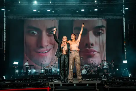 Ca7riel and Paco Amoroso, arms up, in front of their giant faces on the screens. Granca Live Fest, Gran Canaria, July 2025. Photo for Why Agency.