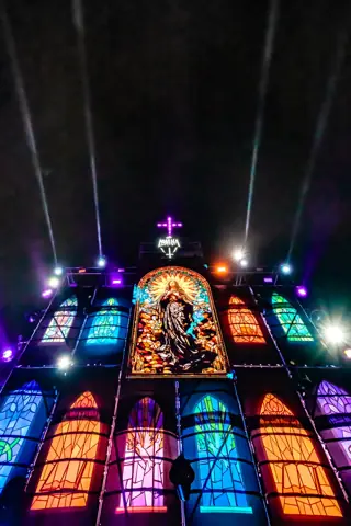 Fachada de vidrieras iluminadas con una Virgen y haces de luz en la noche. La Misa, Gran Canaria, marzo 2024