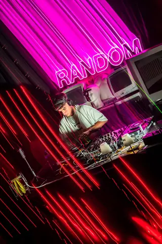 Rayconen pincha bajo un neón rosa de Random, con estelas de luz roja. Rayconen, mayo 2025