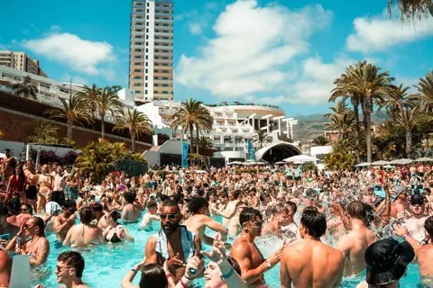 Fiesta en la piscina con agua salpicando, palmeras y hoteles al sol. Cru2, Tenerife, junio 2026