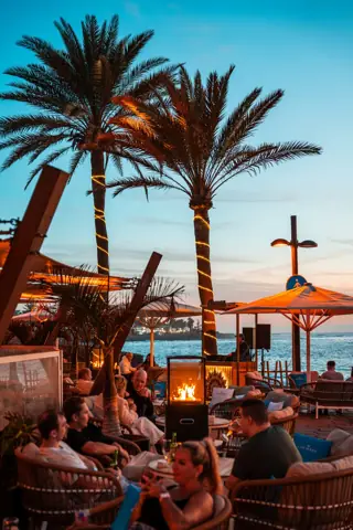 Terraza con palmeras iluminadas, fuego y DJ frente al mar al atardecer. Papagayo Beach Club, Tenerife, febrero 2026