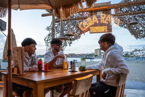 Tres amigos con cócteles bajo el neón de Casa Playa frente a la playa. Red Bull × Casa Playa, Tenerife, diciembre 2025